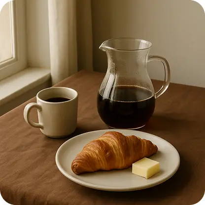 Breakfast setup with a cup of coffee, a jug of brewed coffee, a croissant, and butter on a plate